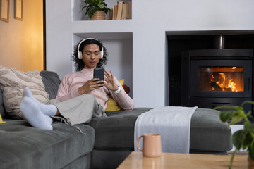 Naklejka premium Nonbinary adult reclining on gray sofa at home wearing headset holding phone fireplace glowing