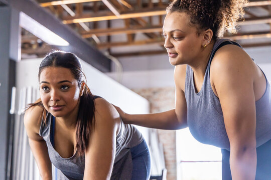 Diverse female trainer in athletic wear guiding client doing bent-over move in gym with beams