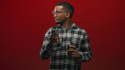 Obraz premium Man with glasses in plaid shirt looking down at smartphone while holding a paper coffee cup against a red studio backdrop; concentration morning routine.