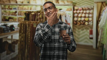 Fototapeta premium Young black man wearing plaid shirt and glasses holding coffee cup and touching chin in boutique building; thoughtful reflection.