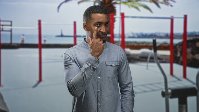 Man points index finger to eye in gray shirt beside red pullup bars and seaside pier at street outdoor gym; quiet confidence.