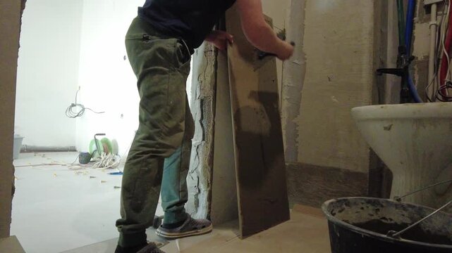 A man is applying cement to tiles in an apartment as part of a renovation. The tools and materials used for the job are visible throughout the room. Dust and unfinished areas are present