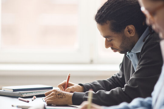 Multiethnic young adult man writing notes during classroom session, showing focus, learning, academic engagement. Useful for education marketing, training programs, student success content