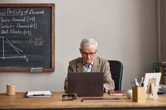 Senior Caucasian male teacher working on laptop beside economics chalkboard, preparing lesson content and online instruction. Useful for education marketing, e learning, academic training