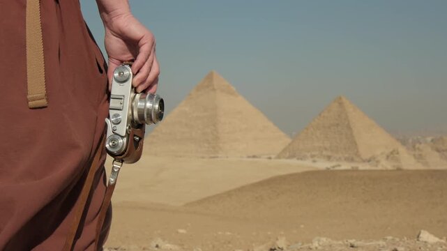 Woman traveler photographer with retro camera in hand pyramids of Giza, wearing pants flapping in wind, while standing on sandy plateau on tourist tour in Cairo Egypt