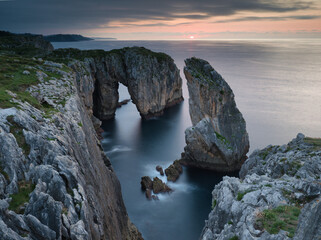 Fototapeta premium Epic Long Exposure Coastal Sea Arch at Sunset - Dramatic high-angle long exposure view of a massive natural sea arch and eroded cliffs framing the sunset over the Atlantic Ocean in Asturias.