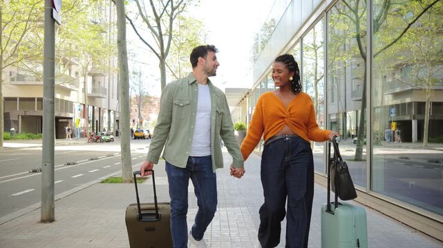 A cheerful couple strolls together down the city street, each holding luggage and refreshments, embodying the spirit of travel and adventure while enjoying each other's company on a sunny day.