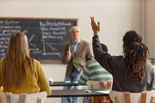 Black teenage boy raising hand during classroom lesson while senior male teacher explaining economics to students. Useful for education marketing, school promotion, student engagement