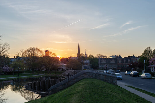 Panoramic sunset view of the historic clustered spires skyline and Carroll Creek in Frederick, Maryland, with a stone bridge and quiet city street.