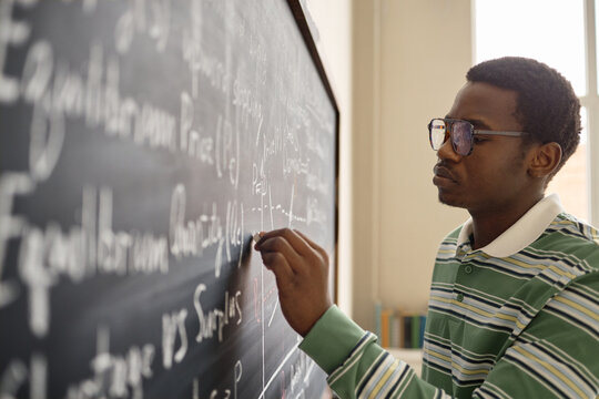 Young African American man writing equations on chalkboard in classroom, studying mathematics and problem solving. Useful for education marketing, tutoring services, STEM learning campaigns