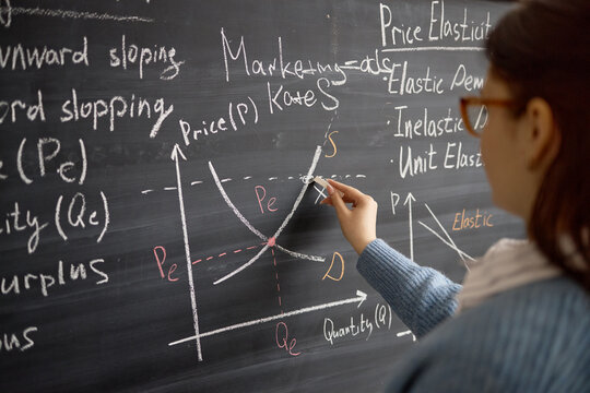Young adult woman writing supply and demand graph on chalkboard during economics lesson, explaining price elasticity concept. Useful for education marketing, tutoring, business training