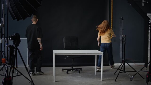 Young Caucasian couple preparing modern studio for video recording, they placing wooden table in center of room and adjusting black backdrop