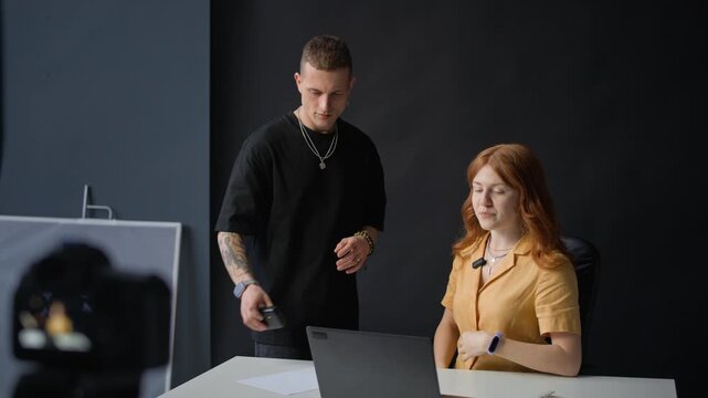 Young Caucasian man coming to woman, putting lav mic on her yellow shirt and starting using laptop in studio