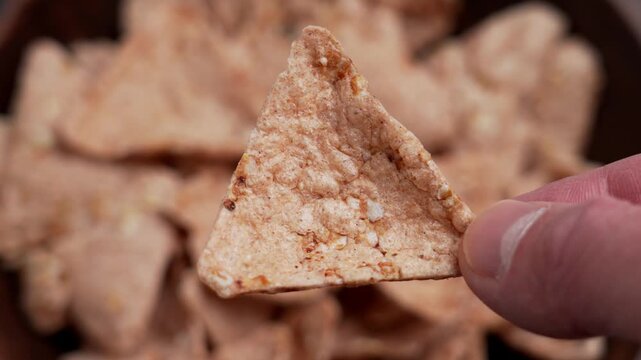 Fingers holding crispy organic buckwheat tortilla chip over a full wooden bowl. Gluten free nachos with grains of amaranth and quinoa