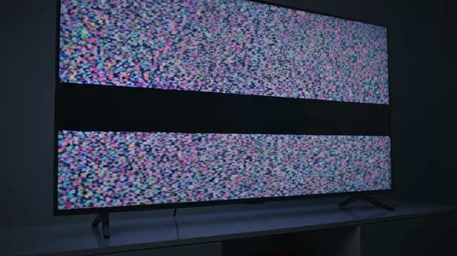 This captures a large screen TV in a dark room displaying technical difficulties with a distorted and glitchy error screen. The screen is filled with colorful static and pixels, indicating a