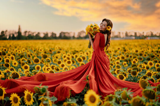 Beautiful smiling woman in red dress holding bouquet of sunflowers standing in blooming sunflower field at sunset, warm summer portrait with golden natural light