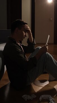 Vertical video young man sitting on the floor in a dimly lit room, holding a letter, feeling overwhelmed and worried about bad news, financial problems, or a difficult situation