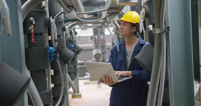 A male technician in a hard hat inspects an HVAC system with a laptop, performing quality control and maintenance checks in an industrial plant.