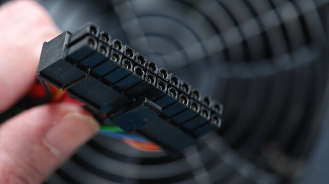 A hand holds a 24-pin ATX power connector with visible dust on its surface against the fan grill of a computer power supply unit. Close up.