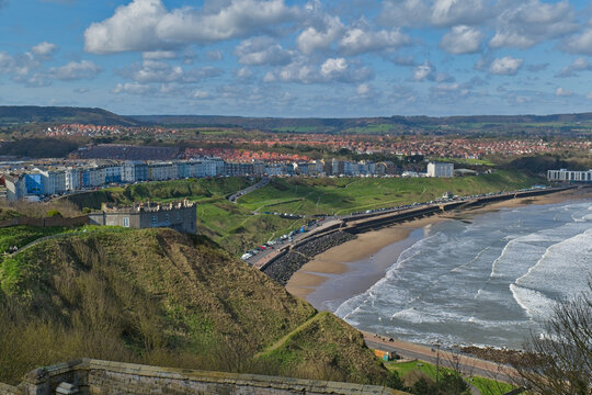 An elevated view captures a coastal town with a sandy beach, breaking waves, and buildings along the cliffs. A fort-like structure sits on a green hill  in Scarborough Castle - Yorkshire - Great Brita