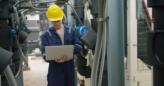 A male technician in a hard hat inspects an HVAC system with a laptop, performing quality control and maintenance checks in an industrial plant.