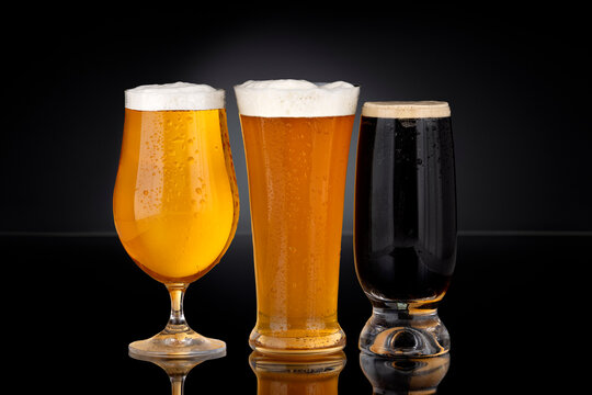 Three glasses of beer including lager, wheat beer and dark stout on black background with foam and condensation. Cold refreshing alcoholic beverages in commercial product photography style.