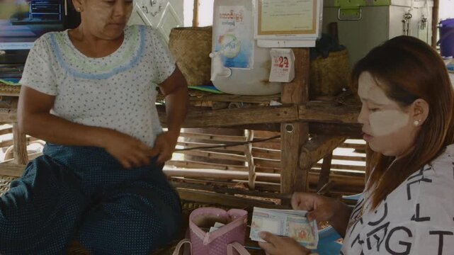 Asian woman counting Myanmar kyat banknotes and giving them to another woman inside a bamboo hut