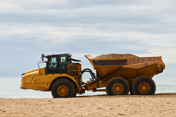 Obraz premium Heavy dump truck driving along sandy beach near the sea, transporting sand and shaping shoreline. Industrial work on coastal area under cloudy sky.
