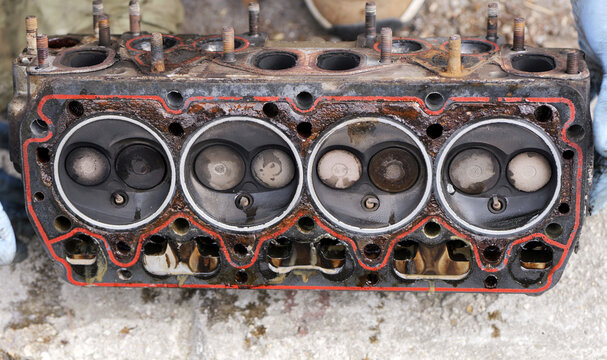 Face of a four-cylinder engine cylinder head from a 1990s car placed upside down on concrete, showing combustion chambers, valves, and worn gasket with deposits. 2026