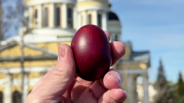 Hand holding a red Easter egg with a blurred church building in the background. Concept of Easter celebration, spring and traditional symbolism