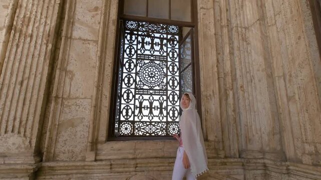A female tourist stands by an ornate window inside a historic mosque in Cairo, Egypt, captured in slow motion 4K during travel.
