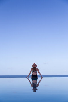 Rear view of a young Woman in a swimming costume and straw hat sitting on the edge of an infinity swimming pool
