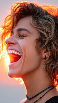 A young man with curly hair laughs heartily with his eyes closed, bathed in the warm, orange light of a sunset