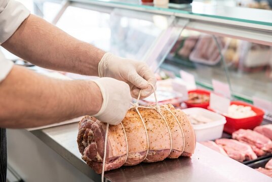 Butcher tying a rolled raw pork roast with butcher twine on a stainless steel meat counter