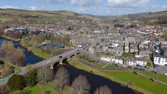 A stunning focal view of the historic town of Peebles, centered on the iconic church spire and the ancient stone bridge spanning the River Tweed. The composition highlights the rich architectural heri