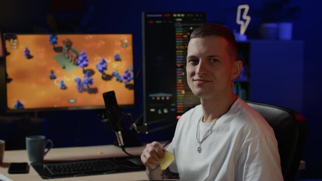Medium shot of young adult Caucasian streamer sitting in computer chair, holding chips, turning to camera slowly and breaking into smile