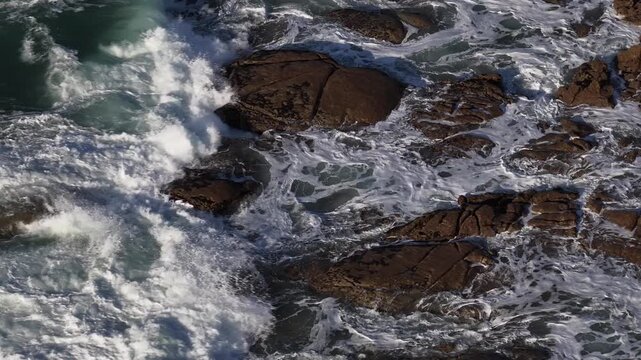 Aerial view of the churning ocean waters crashing against the dark, jagged rocks, creating a dynamic and contrasting seascape, Vila Praia de Ancora, Portugal.