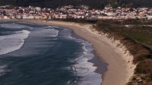 Aerial view of the beach where foamy waves meet the sandy shore, contrasting with the backdrop of the town's buildings, Vila Praia de Ancora, Portugal.