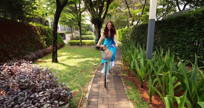 Young woman rides her bicycle along narrow path in residential park, handlebars basket occupied by charming French Bulldog puppy. Slow-motion shot of carefree and easy ride in warm summer weather.
