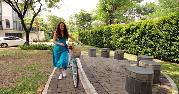 Woman leisurely strolls along narrow pathway in suburban park, pushing her bicycle with cute puppy in handlebar basket. Carefree and relaxed atmosphere on summer day, slow-motion, reverse dolly shot