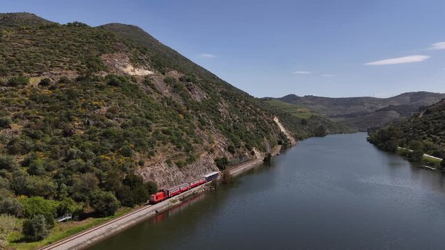 Aerial view of the striking red train contrasting against the green hills and blue river under the clear sky, offering a scenic journey, Regua, Portugal.