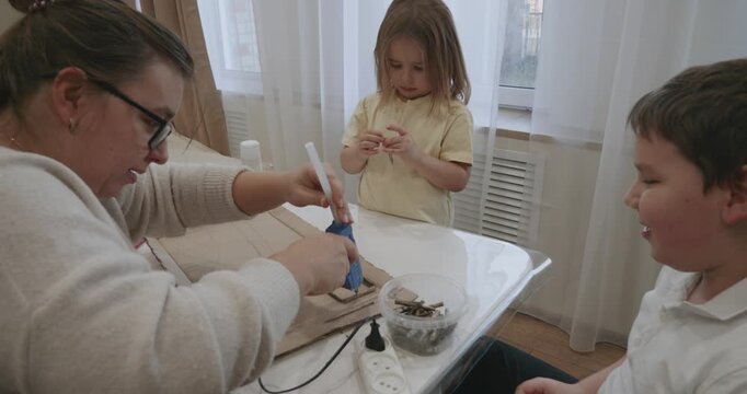 Creative family doing needlework together at the table. A mother uses a hot glue gun to assemble a project from cardboard and twigs while her son and daughter help. The family is making a craft.