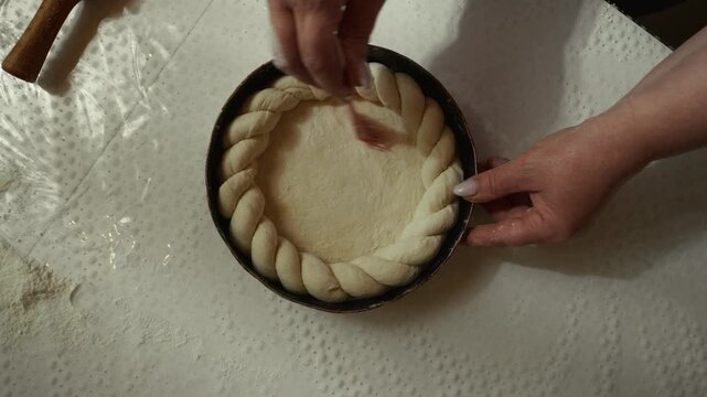 Female baker prepares braided dough in round baking pan on white countertop, with rolling pin and flour scattered around, creating a cozy kitchen atmosphere