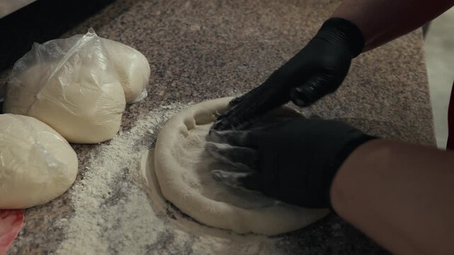 Individual in black gloves shapes dough on a granite countertop, with additional dough balls and flour scattered around, creating a pizza base in a kitchen setting