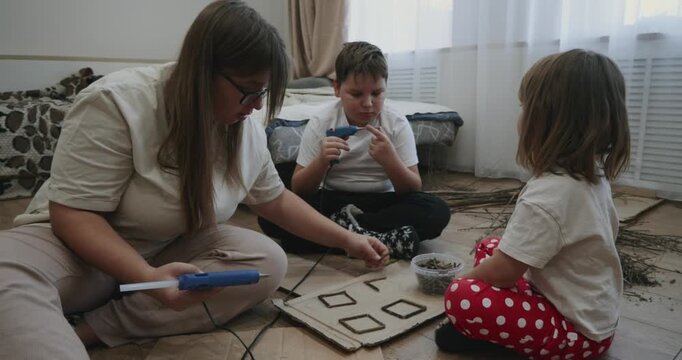 Loving mother helping her son and daughter with a creative school project, using a hot glue gun to attach twigs to a piece of cardboard while sitting together on the bedroom floor.