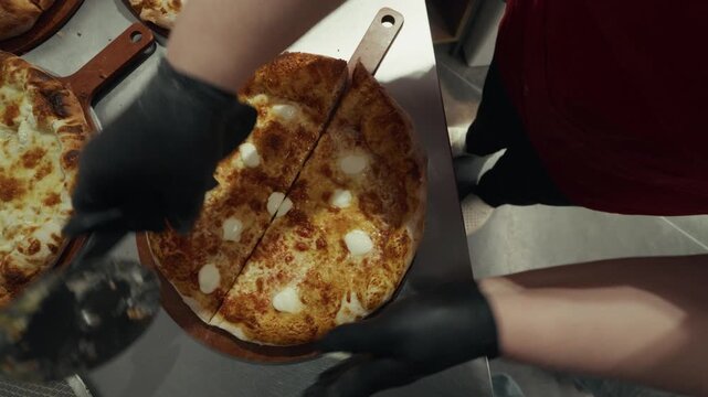 Person in black gloves slices freshly baked pizza with a pizza cutter on a stainless steel countertop, multiple pizzas visible in the background