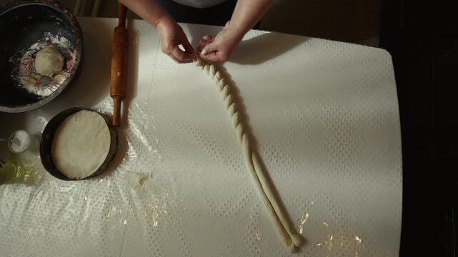 Woman with light skin prepares braided dough on a clean table, using hands to twist and shape the dough while kitchen tools and ingredients are visible in the background