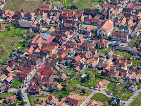 Aerial view of terracotta rooftops nestled amongst verdant trees, the church steeple piercing the skyline, casting shadows over the quaint village, Kintzheim, Grand Est, France.