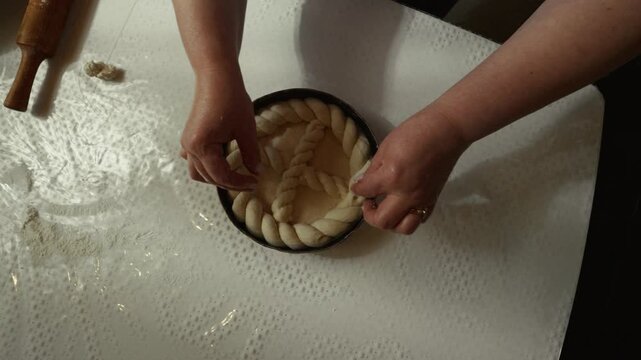 Female baker skillfully shapes dough into a braided pattern inside a kitchen, with a rolling pin and flour-dusted surface visible in the background