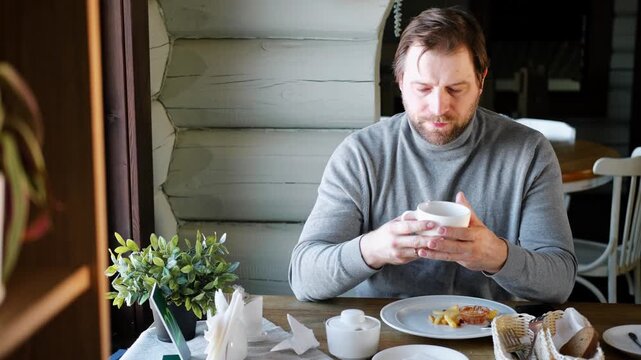 Handsome bearded man has lunch sitting at the kitchen table at home. Male eating French fries potatoes meal from white plate, drinking tea. Happy athlete male enjoying Tasty yummy unhealthy food.